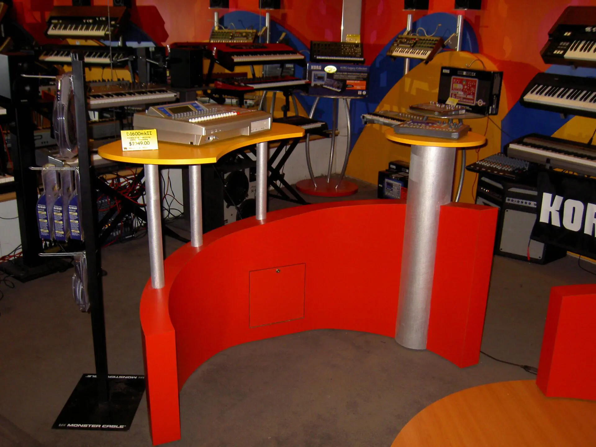 A brightly lit music store display featuring multiple electronic keyboard synthesizers on colorful stands. In the foreground, a vibrant red and yellow curved table showcases two sound mixing consoles. Background shelves are filled with various musical equipment.